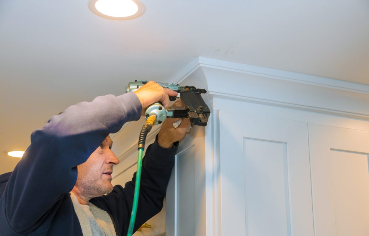 Air nailer tool close up of carpenter using nail gun to crown moldings on kitchen cabinets with white cabinets