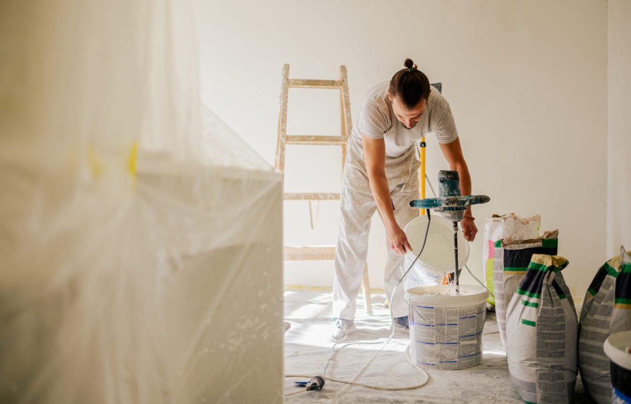 A constructor is standing in a house in a renovation process and adding water to a plaster mixture and preparing for skim coating. A worker is mixing materials in a bucket and preparing for plastering